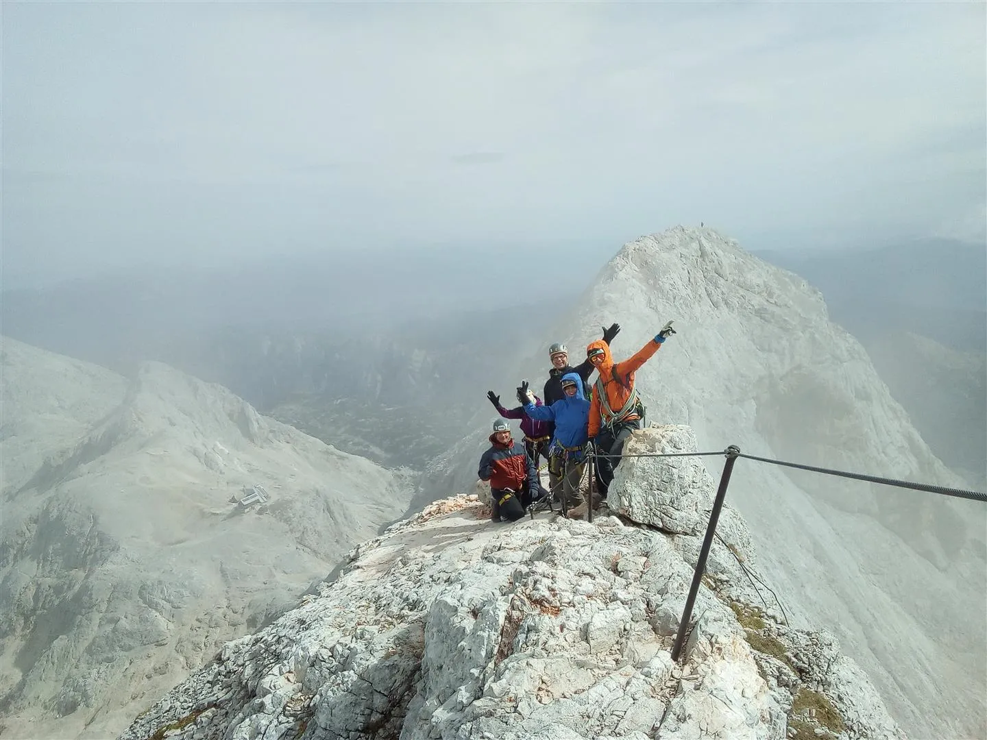 A group of cheerful mountaineers poses on a rugged rocky peak, waving their hands along a misty mountain ridge secured with a steel safety cable.