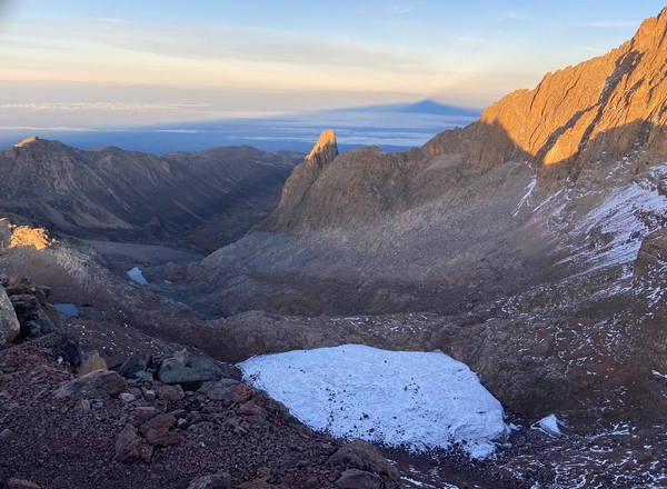 The golden morning sun illuminates the rugged ridges of Mount Kenya, casting a massive triangular shadow over a blanket of clouds in the distant valley.