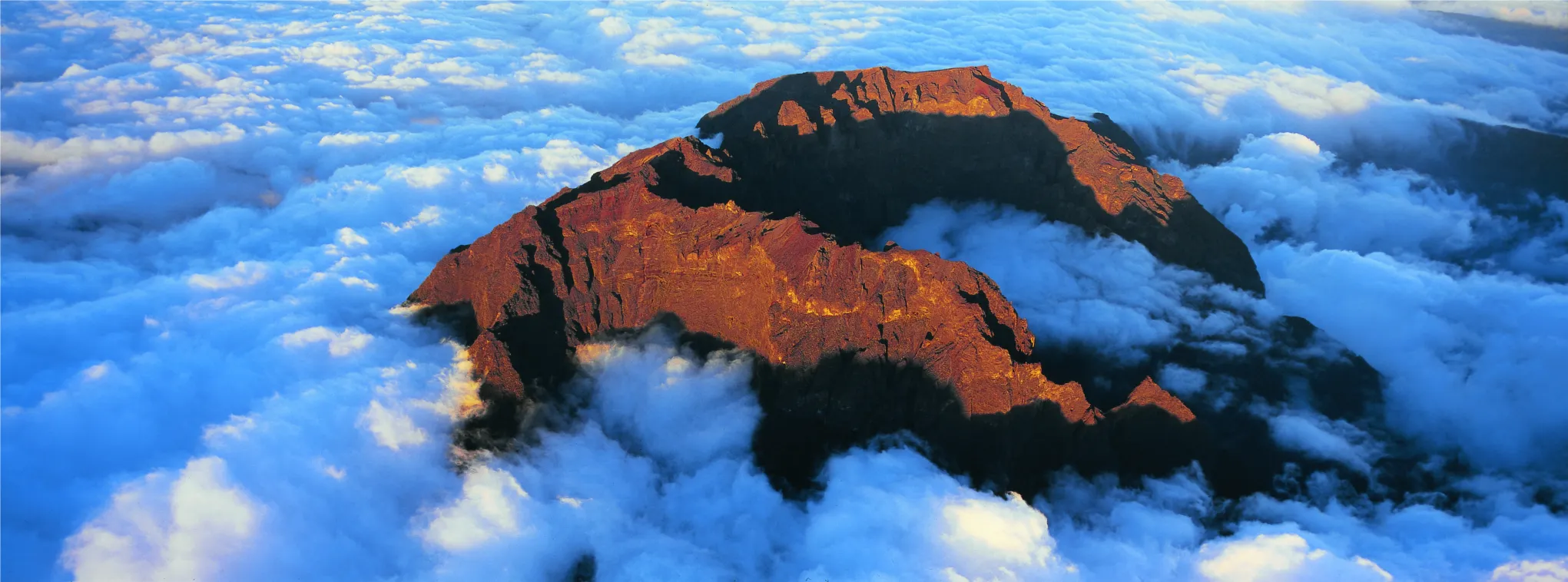 A dramatic aerial view of a rugged volcanic crater glowing in the warm golden light of sunrise, rising above a vast, thick sea of white clouds.