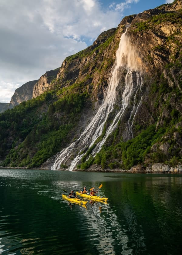 Two yellow kayaks with people kayaking in a fjord below a large waterfall cascading down a steep, green mountain.
