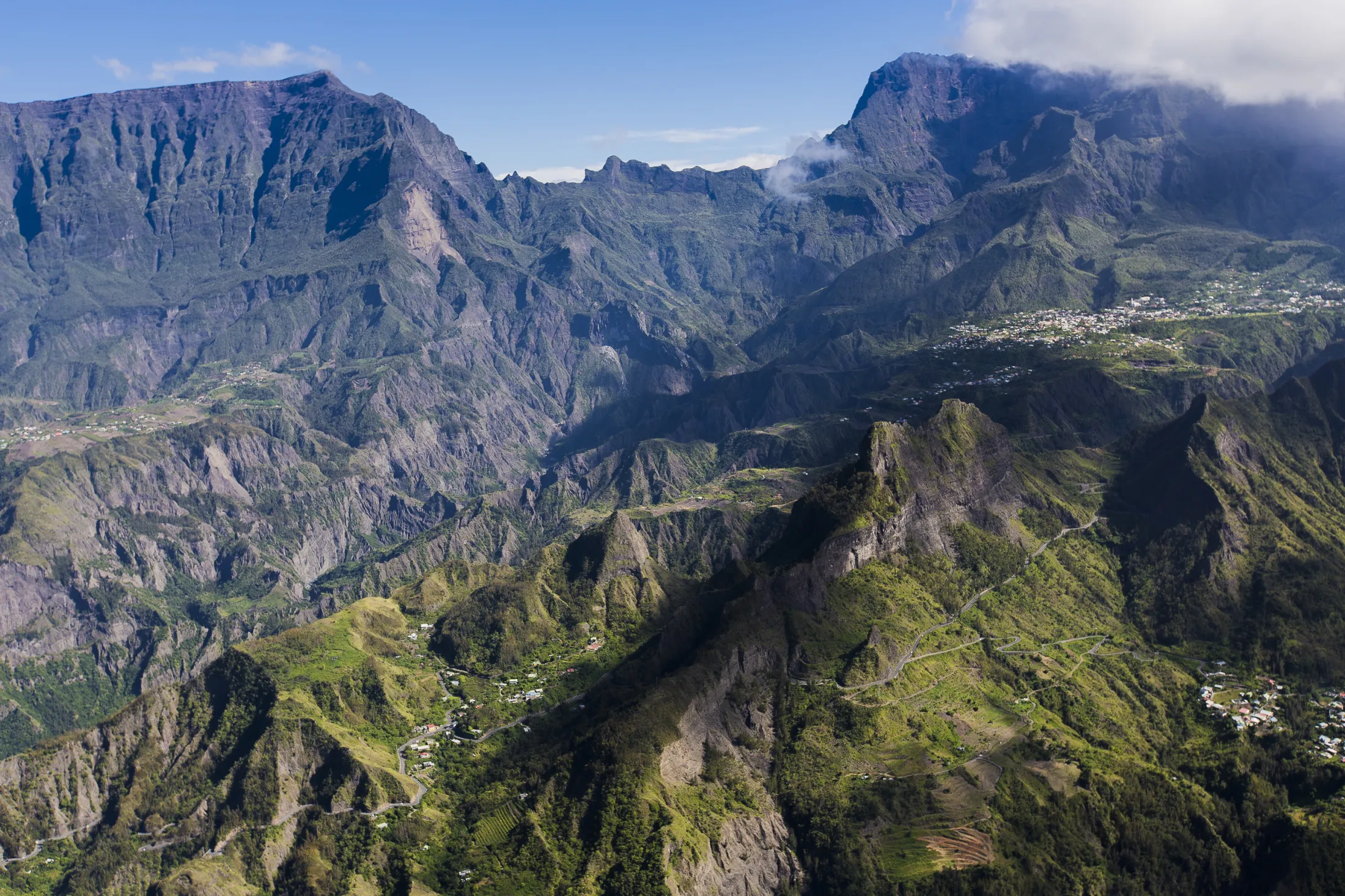 An expansive high-angle view of a dramatic, rugged mountain caldera featuring sharp peaks, deep green valleys, winding roads, and small villages nestled on the slopes.