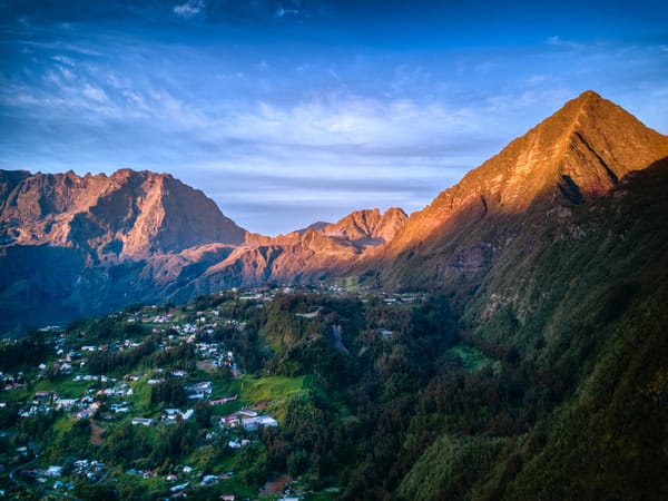 A picturesque mountain village nestled on green slopes under towering volcanic peaks glowing in the warm light of sunrise.