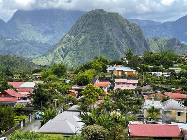 A colorful village with red-roofed houses is nestled among lush tropical vegetation, with a massive dome-shaped green mountain rising dramatically in the background.