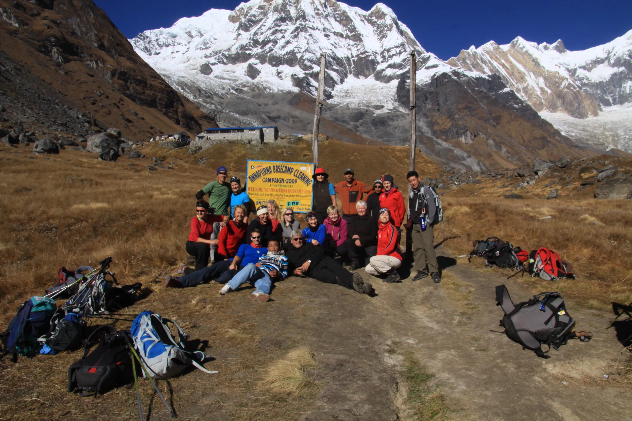A large group of hikers poses together on a dirt trail in front of a yellow Annapurna Base Camp sign, with a dramatic backdrop of towering, snow-capped mountains.