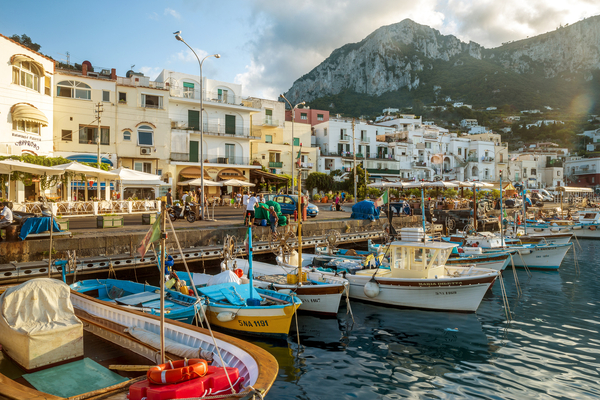 A bustling Italian coastal town harbor with small boats docked against a backdrop of white buildings and a steep mountain.