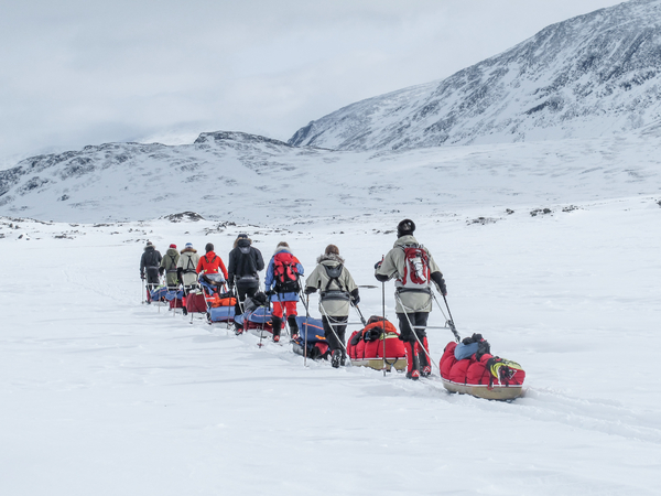 A group of people cross a snowy landscape, pulling sleds with equipment.