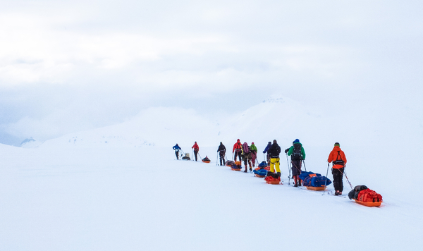 A line of skiers in colorful winter gear pulling sleds across a white, snowy landscape toward distant mountains.