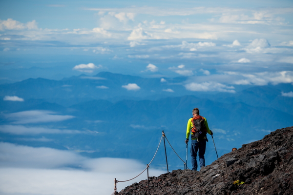 A hiker with a backpack and trekking poles stands on a rocky mountain slope, looking out over a vast landscape of layered blue mountains and clouds.
