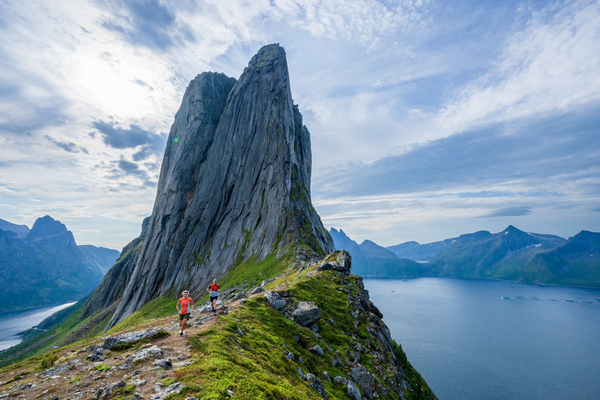 A steep, dramatic mountain peak rises above a fjord with hikers walking along a narrow ridge in the foreground.