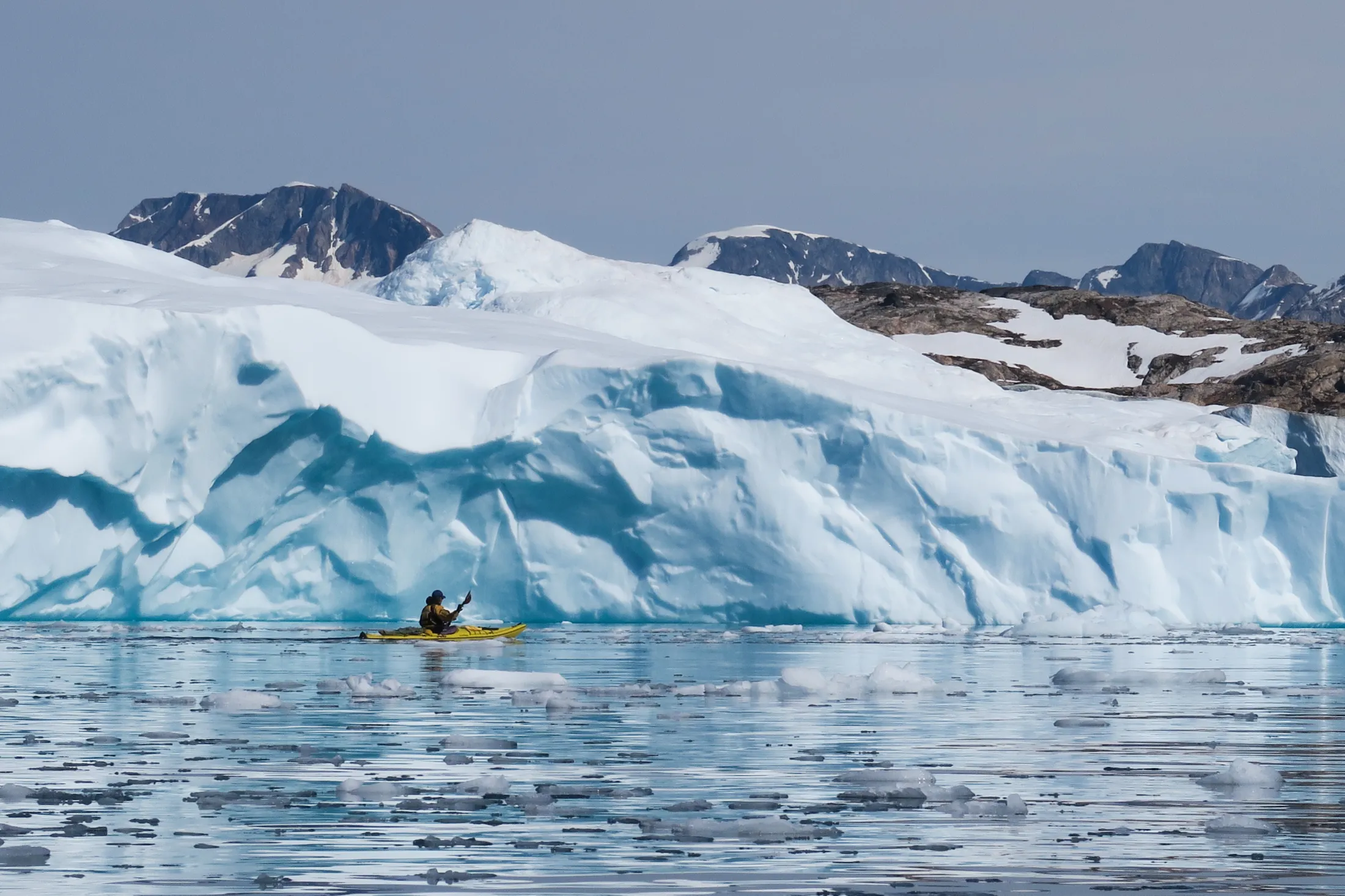A person in a yellow kayak paddles on calm, ice-filled water in front of a massive, light blue ice wall with snow-dusted mountains in the background.