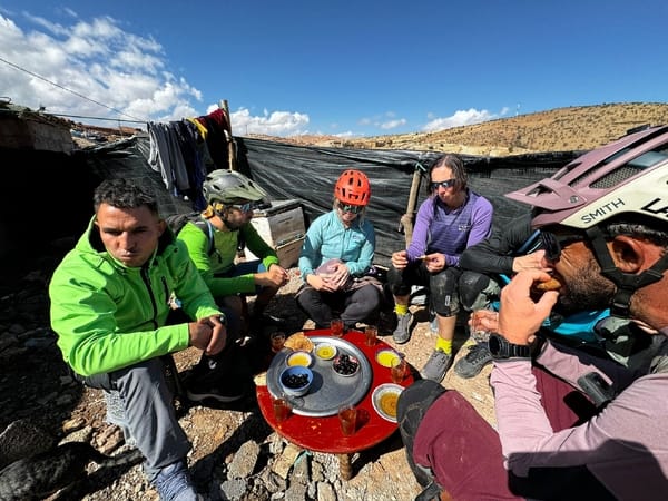 Cyclists eating together outdoors near a low wall under a blue sky.