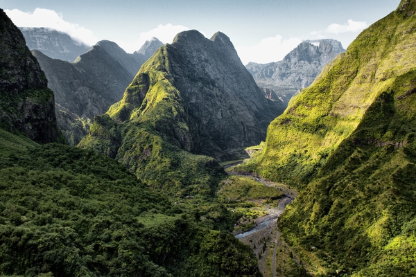 A dramatic view looking down into a deep, lush green mountain valley with a winding river nestled between towering, sunlit volcanic peaks.