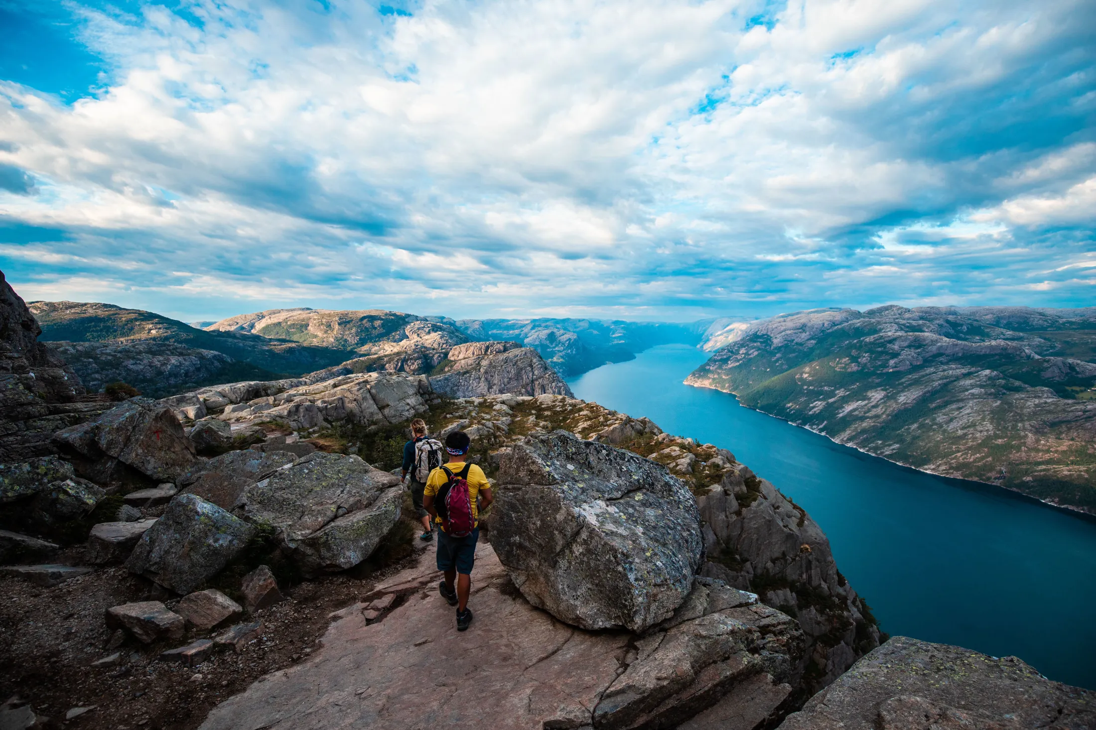 Two hikers in the mountains with the fjord on the right side.