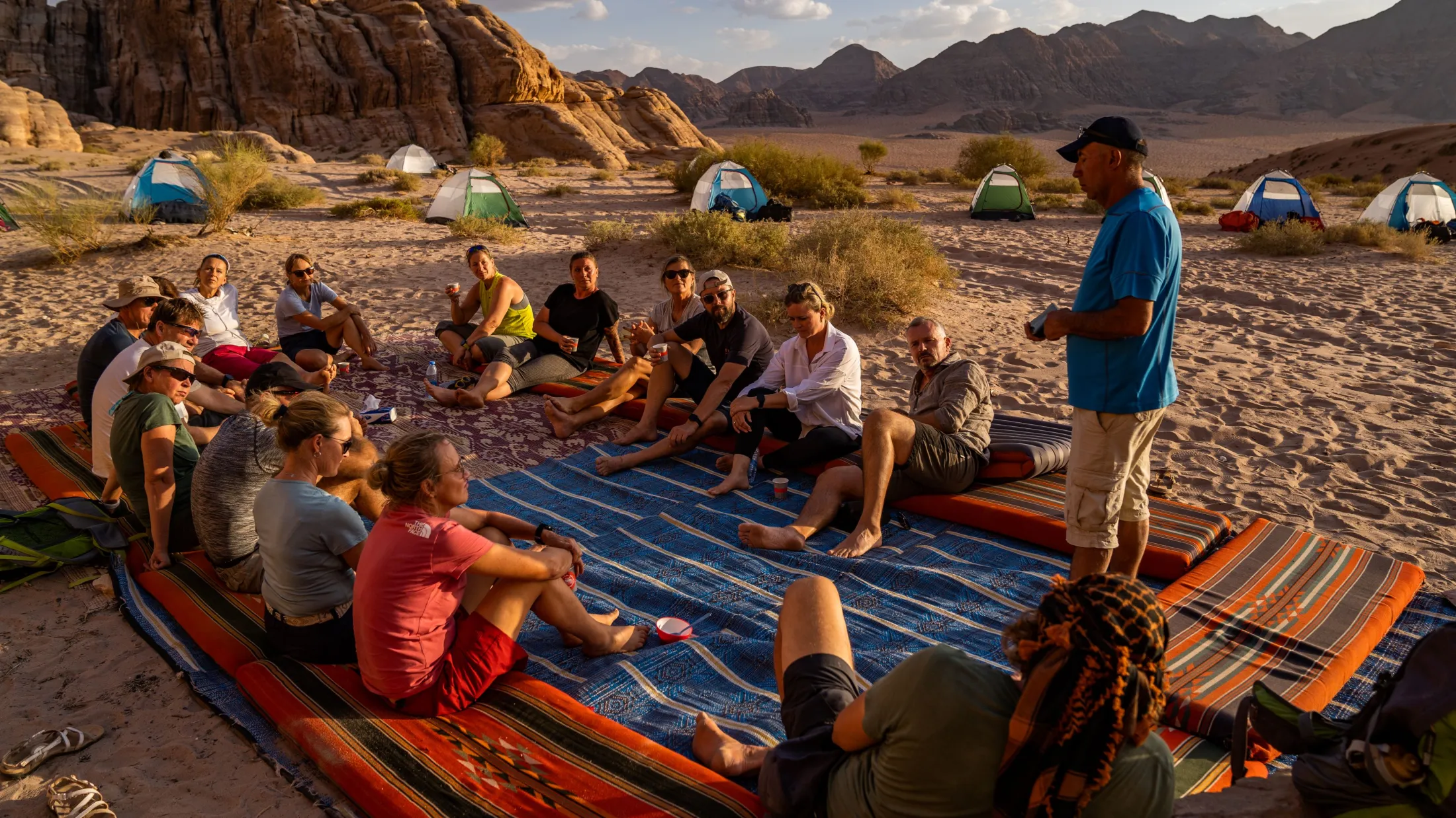 A group of hikers sits together on carpets in the sand at a desert campsite while a man stands and speaks to them.