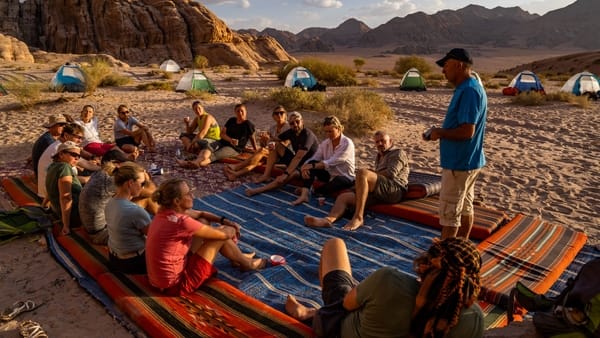 A group of hikers sits together on carpets in the sand at a desert campsite while a man stands and speaks to them.