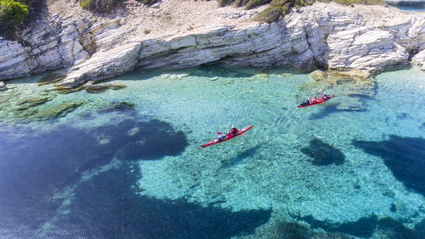 An overhead drone perspective captures red kayaks gliding through remarkably clear turquoise and deep blue water along a rocky, layered limestone shoreline.