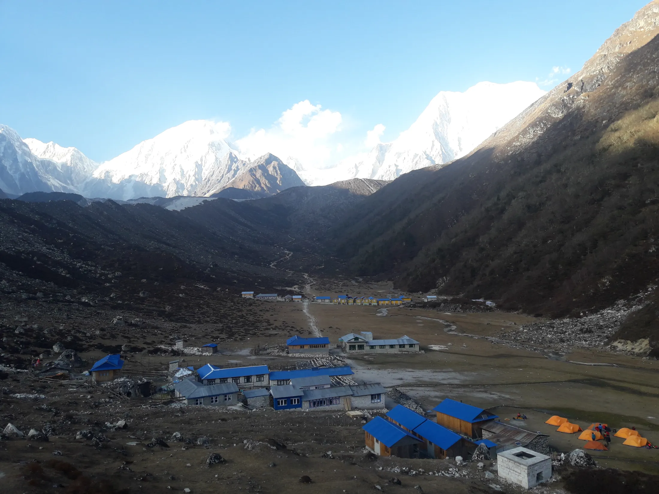English: A remote mountain settlement with bright blue-roofed buildings sits in a vast valley, framed by dark slopes and snow-capped peaks in the background.
