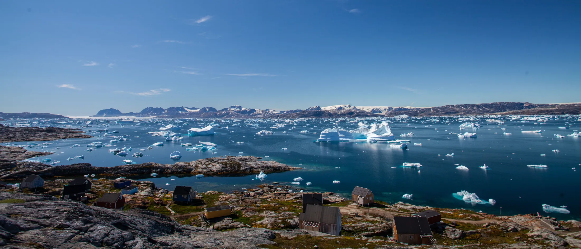 A high-angle view over a small settlement with scattered wooden houses by a deep blue fjord filled with countless icebergs, with snow-dusted mountains and a clear sky in the background.