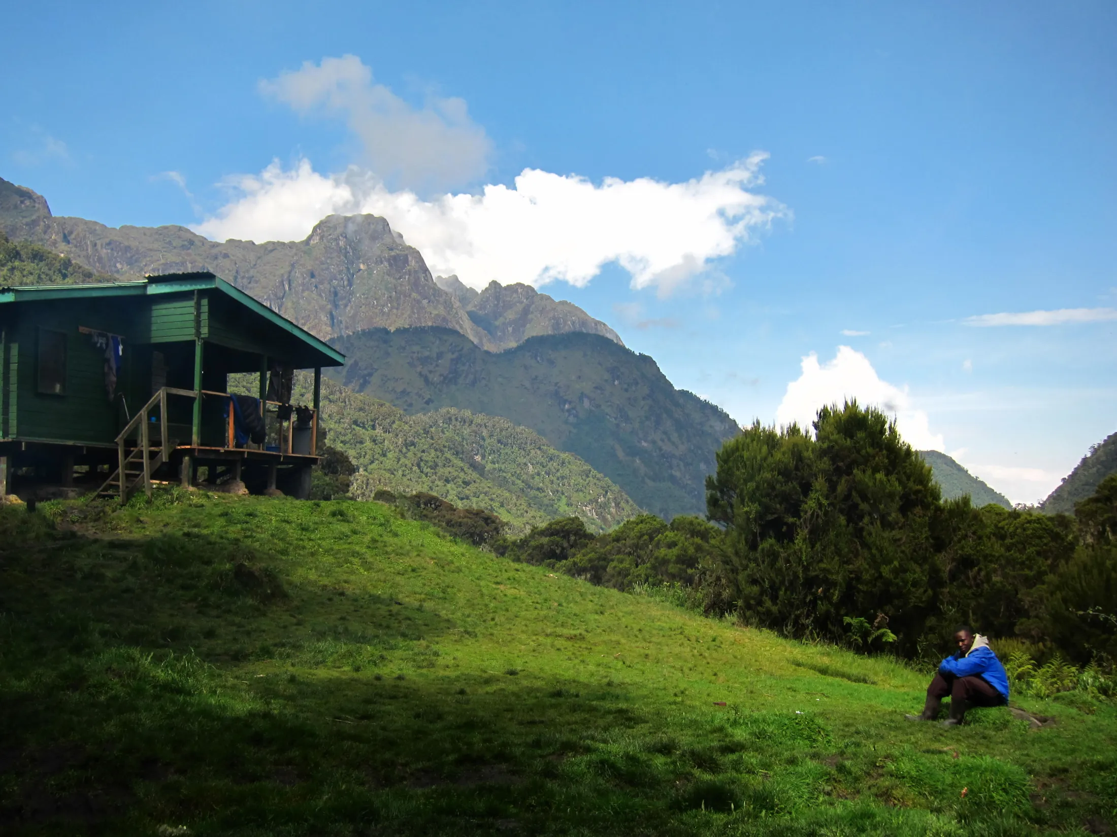 A wooden green mountain hut sits on a grassy slope where a person in a blue jacket sits looking out over a lush forest and towering rocky peaks.