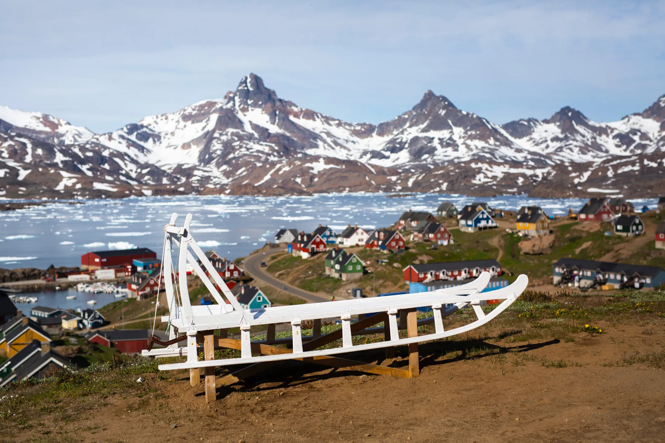 A white dog sled sits on a hill in the foreground overlooking a colorful Greenlandic settlement, with an ice-filled fjord and sharp, snow-covered mountains in the background.