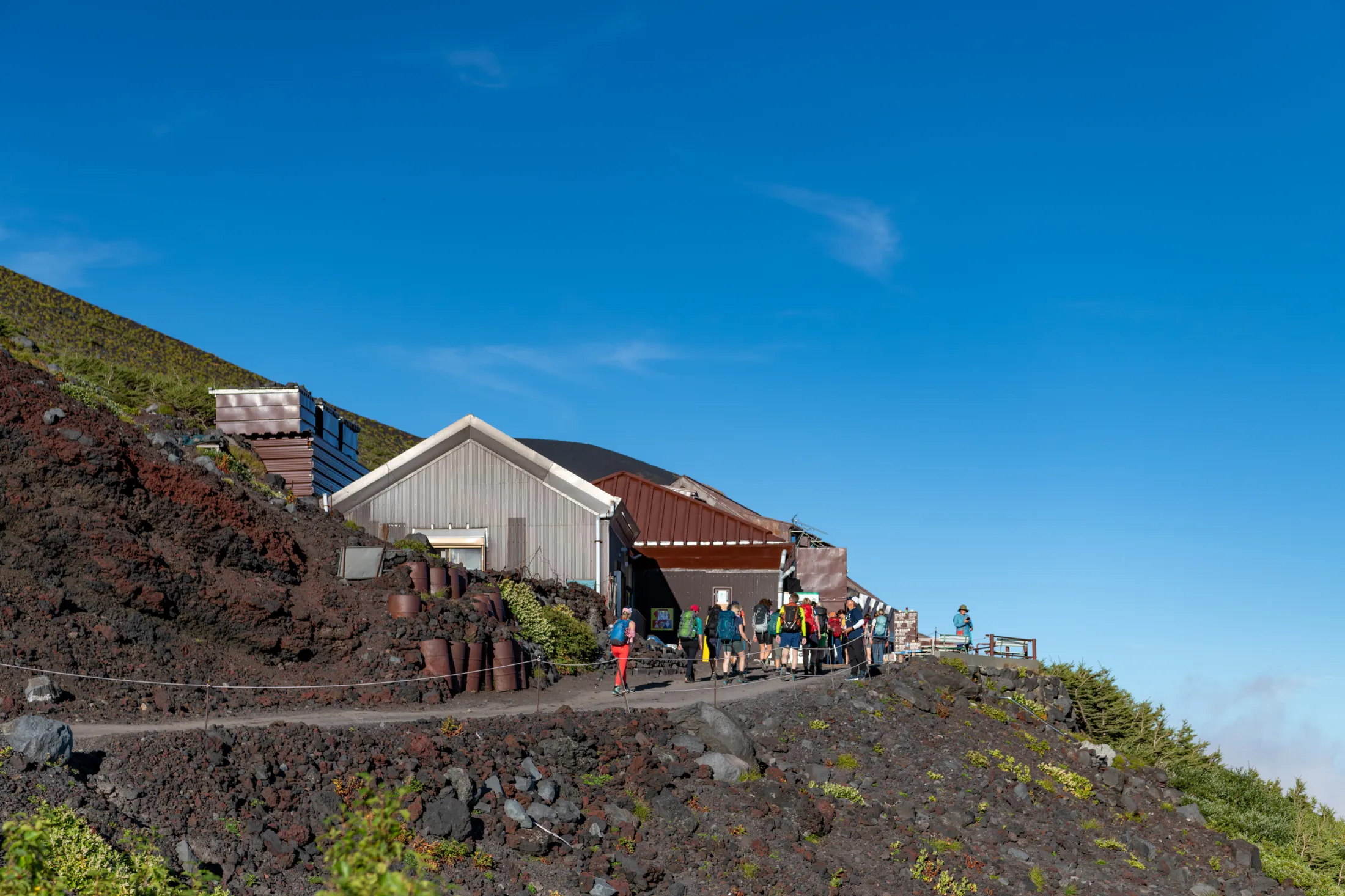 A group of hikers walks along a volcanic, rocky path past simple mountain station buildings under a clear, bright blue sky.