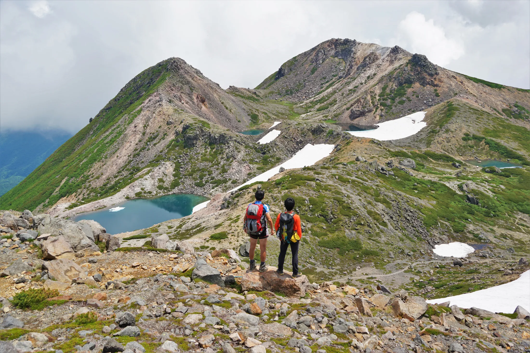 Two hikers with backpacks stand on a rocky overlook, gazing at a dramatic alpine crater landscape with turquoise volcanic lakes and patches of snow nestled between mountain peaks.