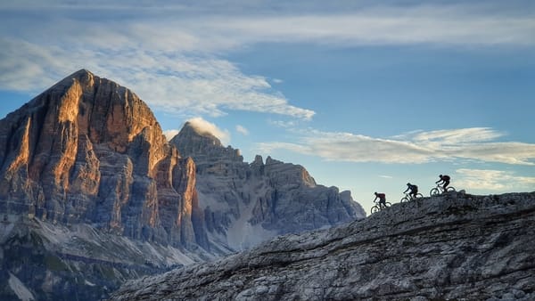 Three mountain bikers riding on a rocky ridge with massive mountain peaks in the background under a blue sky.