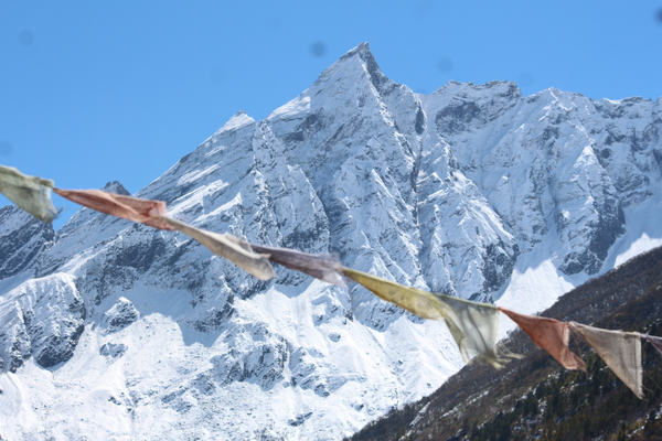 Faded prayer flags flutter in the foreground, framing a sharp, snow-covered mountain peak rising against a clear blue sky.