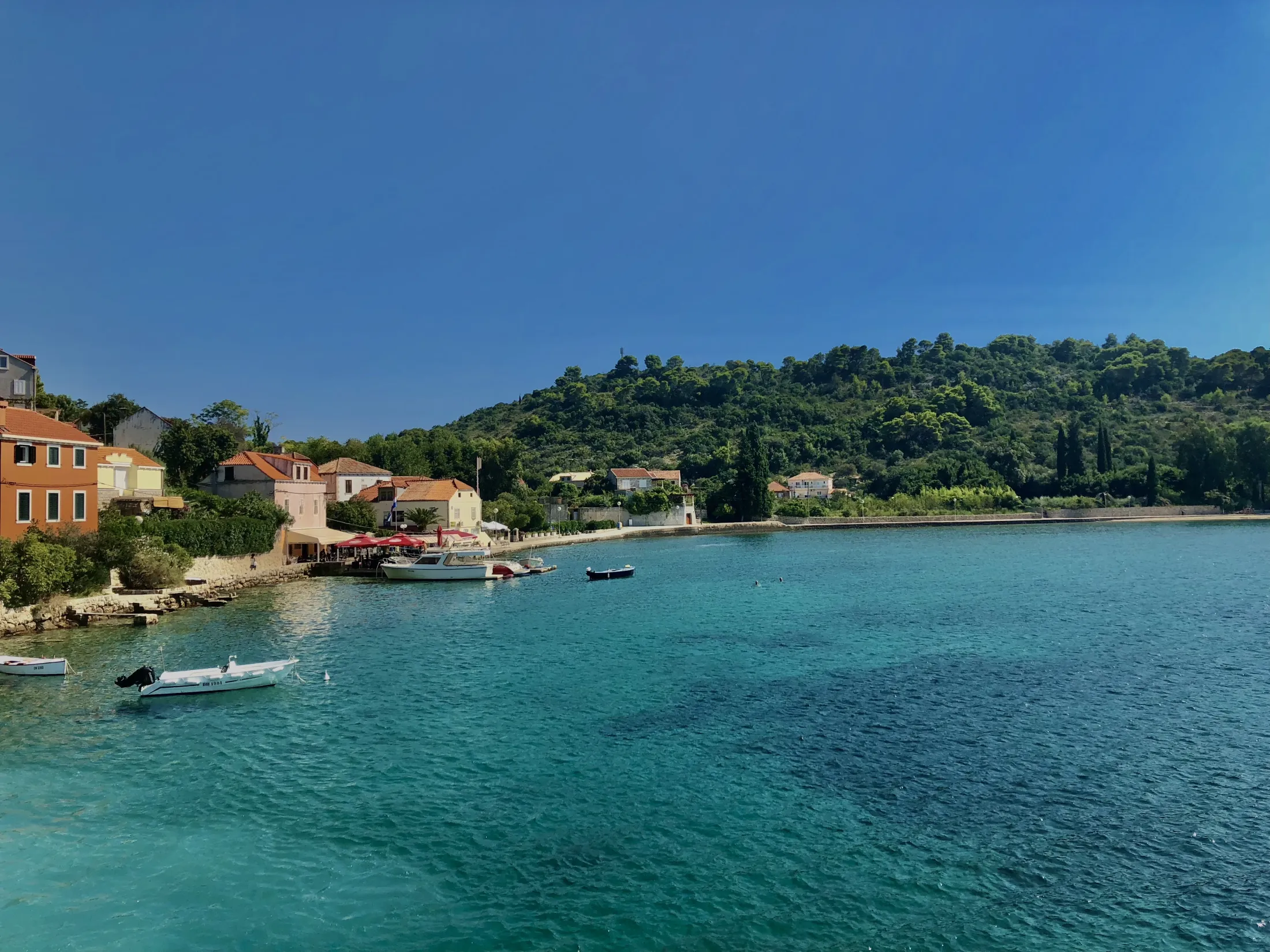A charming coastal village with orange-roofed houses lines a vibrant turquoise bay backed by lush, green hills under a clear blue sky.