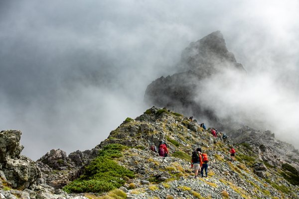 A group of hikers climbs a steep, rocky ridge toward a dramatic, towering peak enveloped in thick, swirling mountain fog.