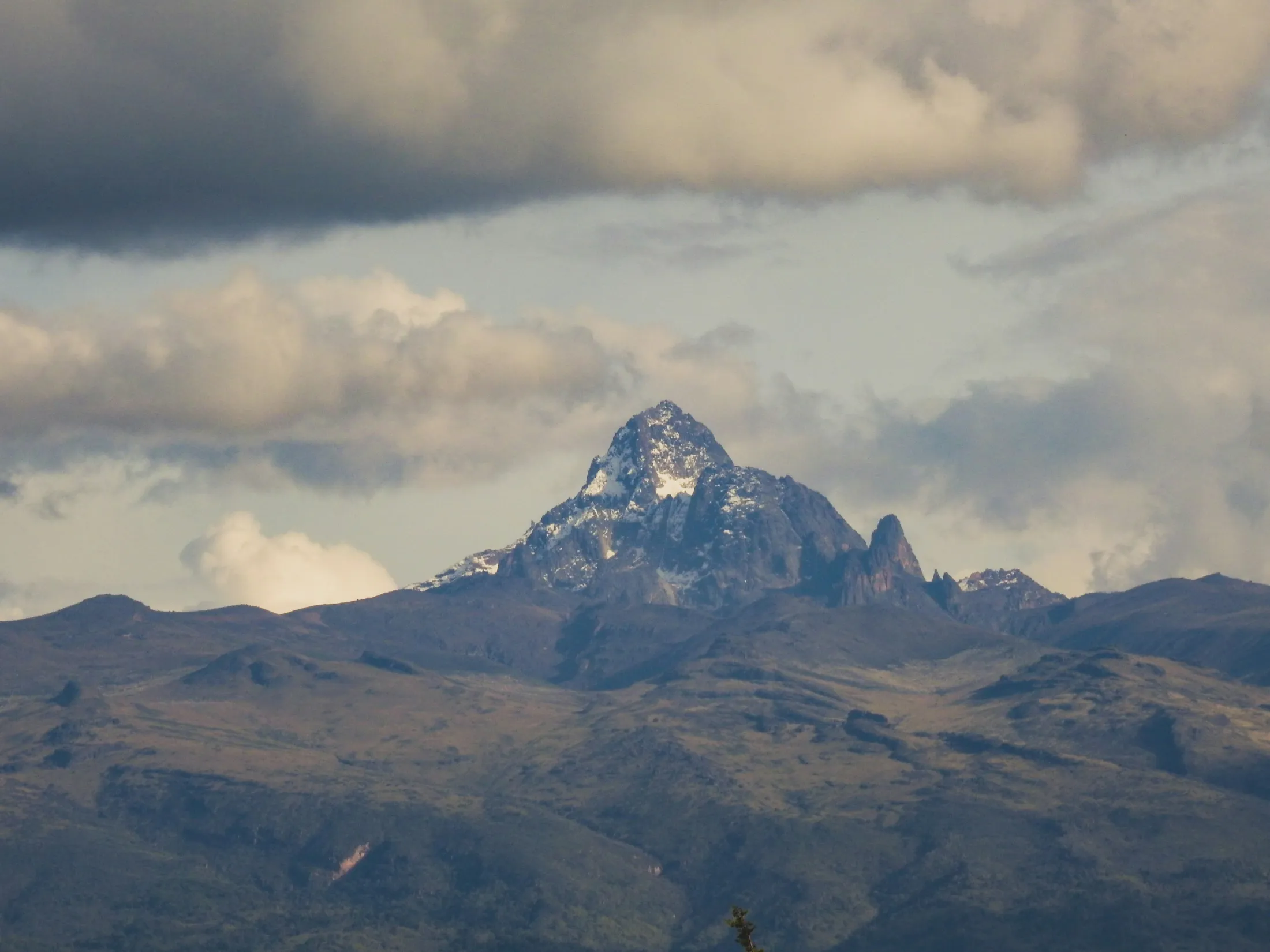 The jagged, snow-dusted peak of Mount Kenya stands prominently beneath a dramatic sky filled with heavy, rolling clouds.