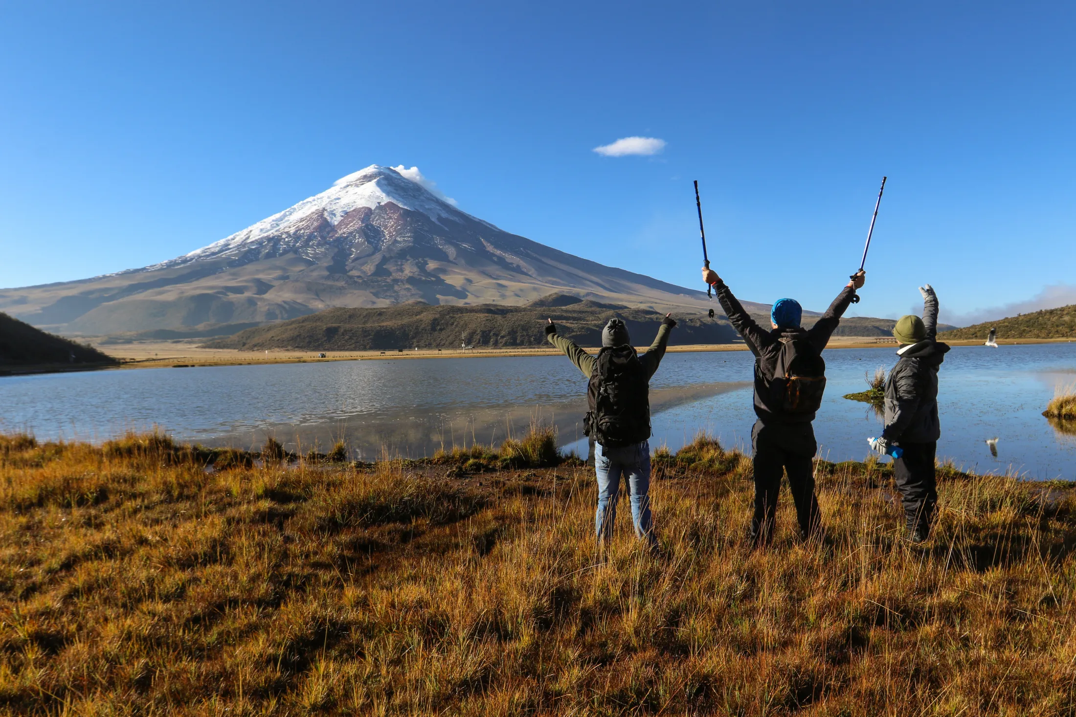 Three hikers stand by a lake, raising their arms in triumph before a snow-capped volcano.