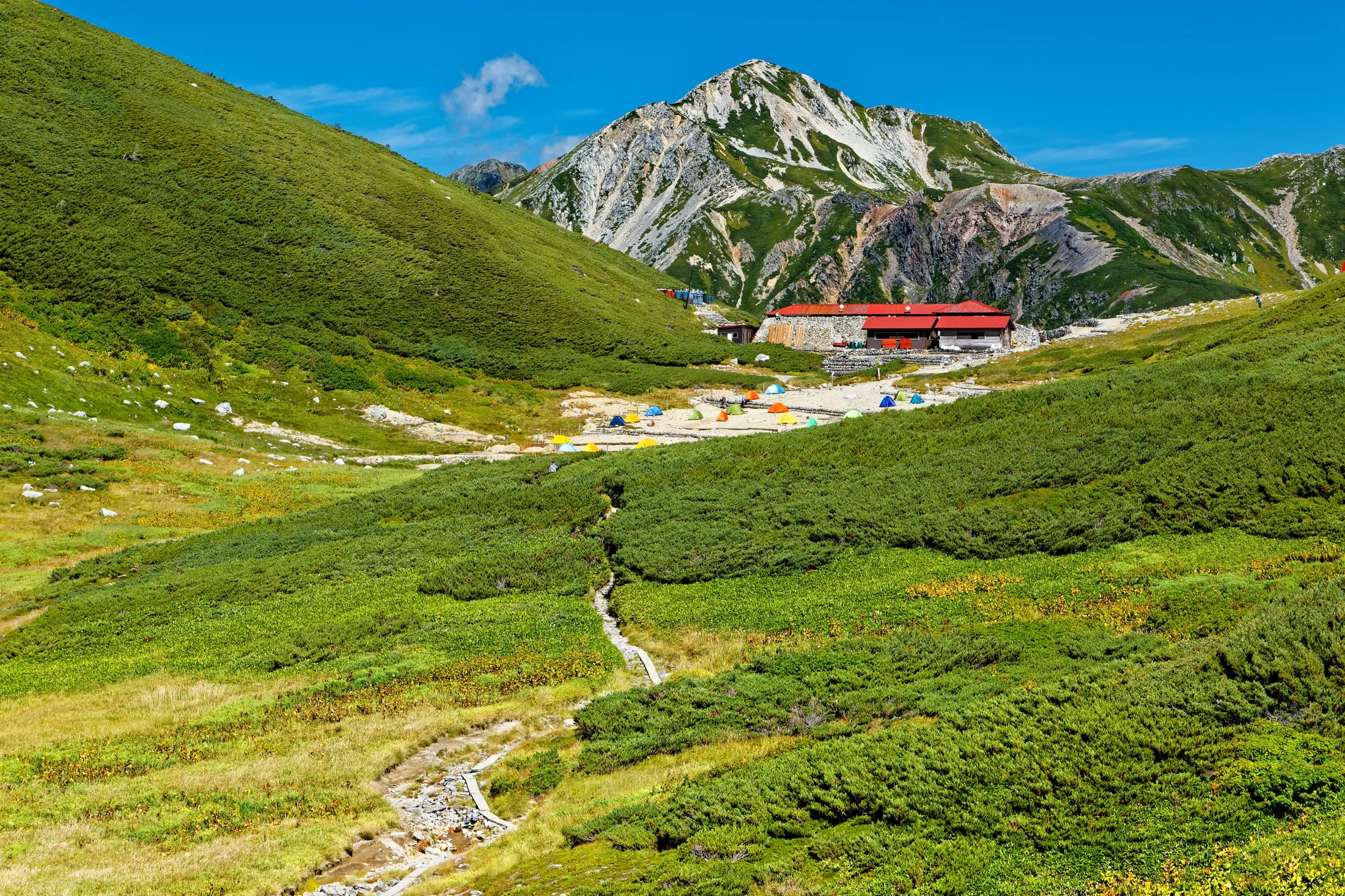 A narrow dirt path winds through lush green, rolling hills toward a mountain lodge with a red roof and a colorful tent campsite nestled at the base of a rugged peak.