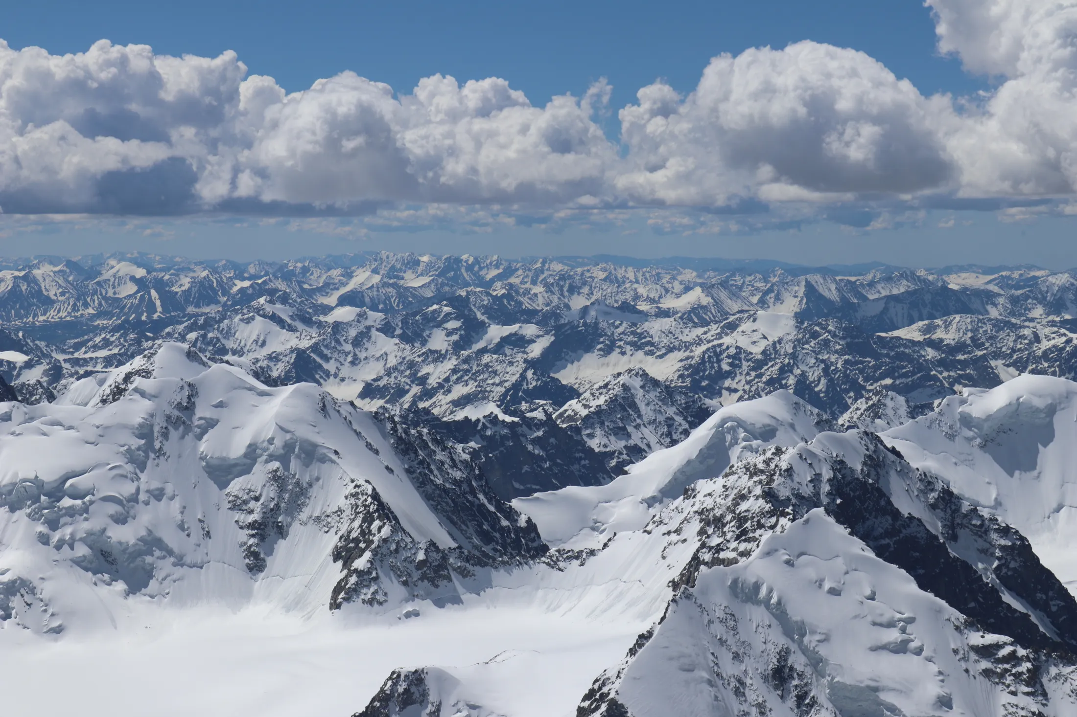 An aerial view looking over a vast, endless sea of sharp, snow-covered mountain peaks stretching to the horizon under a line of puffy white clouds and blue sky.