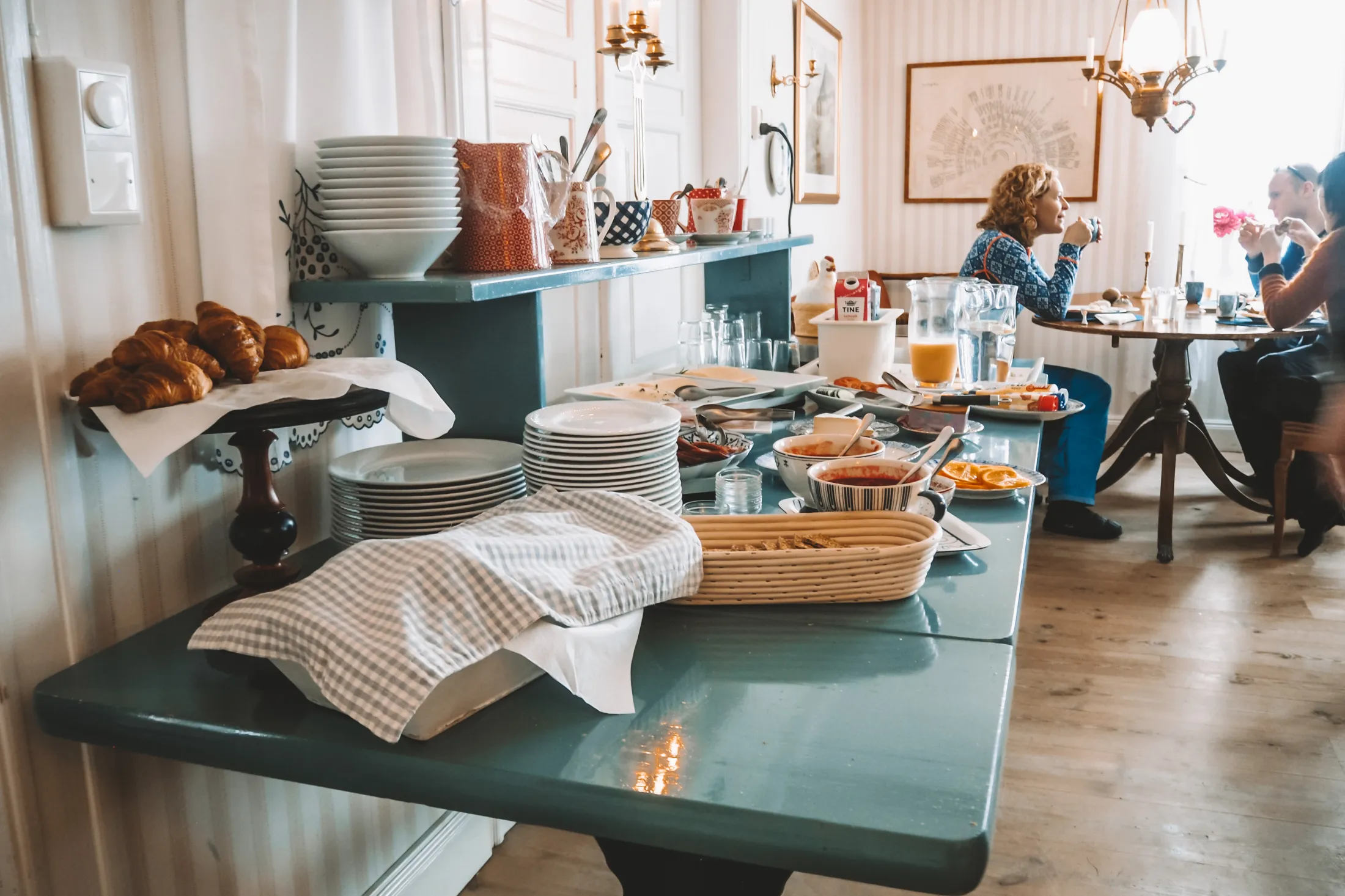 A breakfast buffet table filled with plates, bowls, toppings, and drinks, with a woman sitting and drinking from a cup in the background.