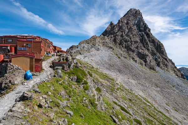 A stone mountain path leads past a collection of wooden alpine huts toward a towering, rugged rock peak under a blue sky with wispy clouds.