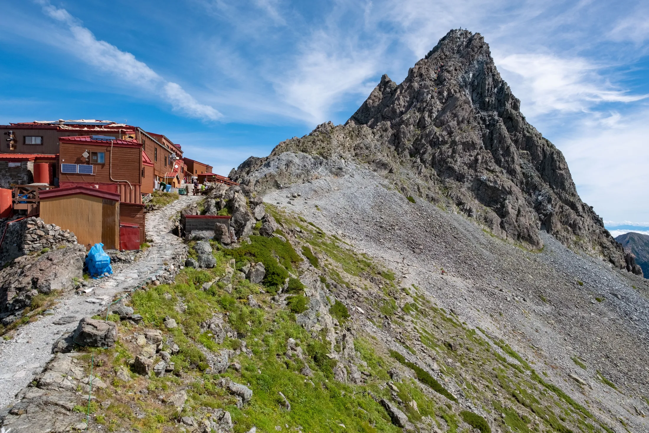 A stone mountain path leads past a collection of wooden alpine huts toward a towering, rugged rock peak under a blue sky with wispy clouds.
