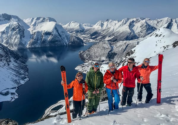Five skiers pose on a snowy mountain ridge with a fjord and snow-covered peaks in the background.