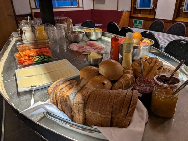A buffet counter laden with breakfast items including sliced bread, rolls, cheese, sliced tomatoes, condiments, and jam.