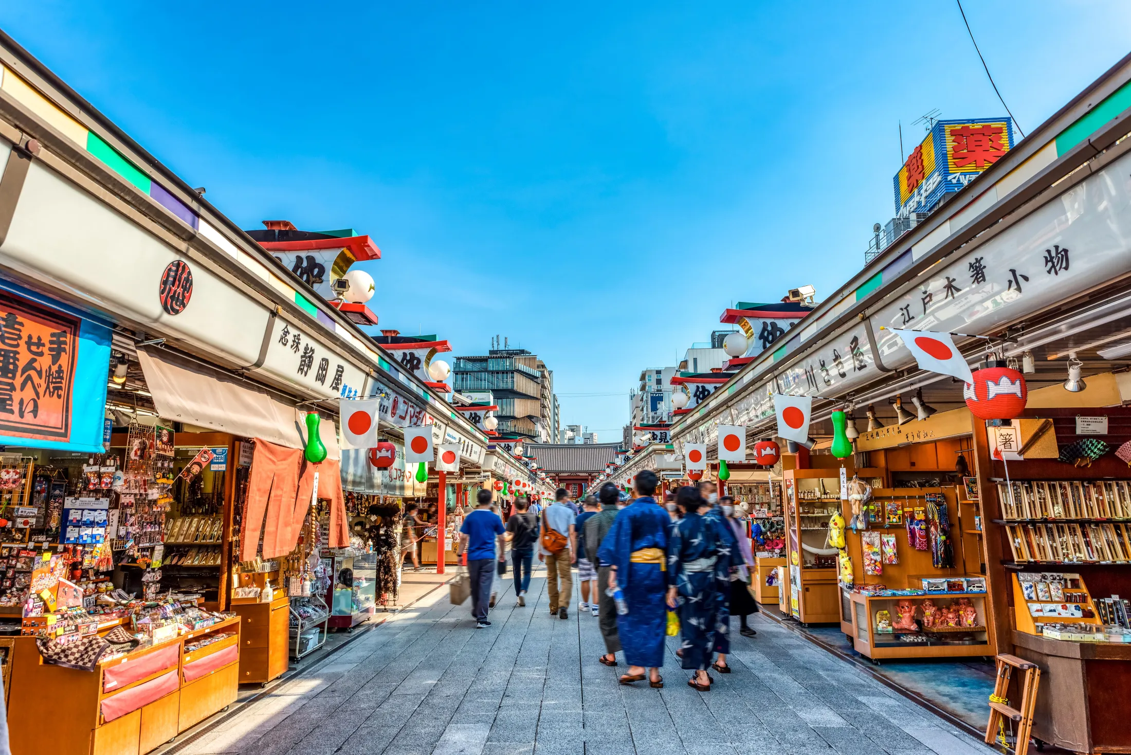 Many shoppers and tourists stroll down a lively, paved market street lined with traditional souvenir stalls decorated with Japanese flags under a bright blue sky.