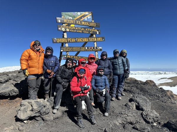 A group of hikers wrapped in warm winter jackets poses together in front of the Uhuru Peak sign on Mount Kilimanjaro under a clear blue sky.