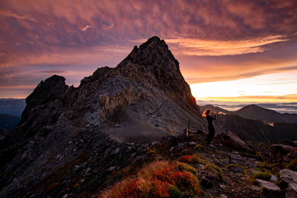A person stands on a rocky mountain ridge taking a photo with their phone during a dramatic, fiery orange and purple sunrise.