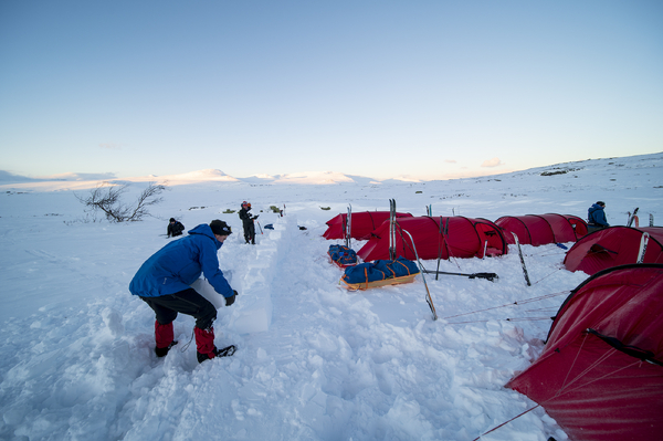 A person placing snow blocks onto a windbreak wall next to a row of red expedition tents in a snowy mountain landscape.