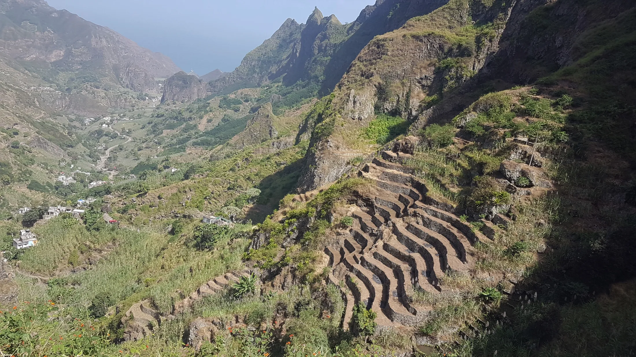 An aerial view of a steep mountain slope terraced with stone walls for farming, surrounded by lush green vegetation and towering, rugged volcanic peaks in the distance.