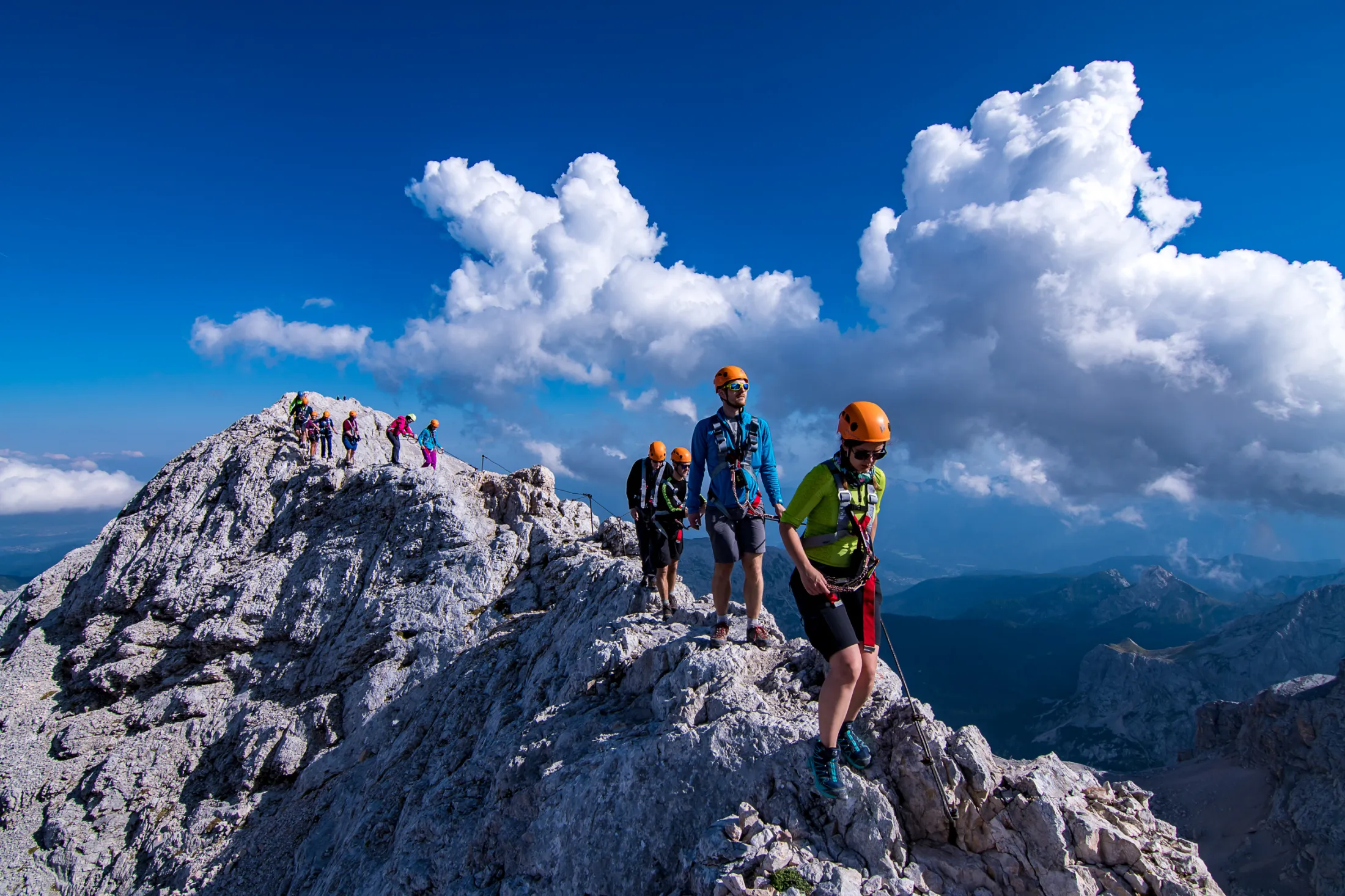A group of climbers wearing helmets and harnesses carefully navigate a narrow, rocky mountain ridge secured by a via ferrata steel cable under a bright blue sky with large white clouds.