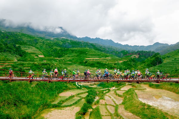 A group of people crossing a suspension bridge over a lush, green valley with terraced fields and mountains in the background.