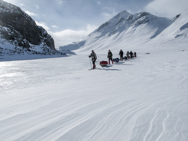 A group of skiers pulling sleds through a snowy mountain valley.