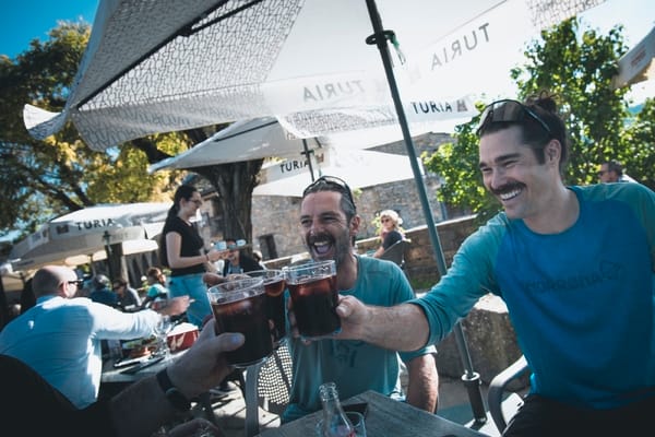 Two men smiling and clinking glasses of dark beer at an outdoor terrace.