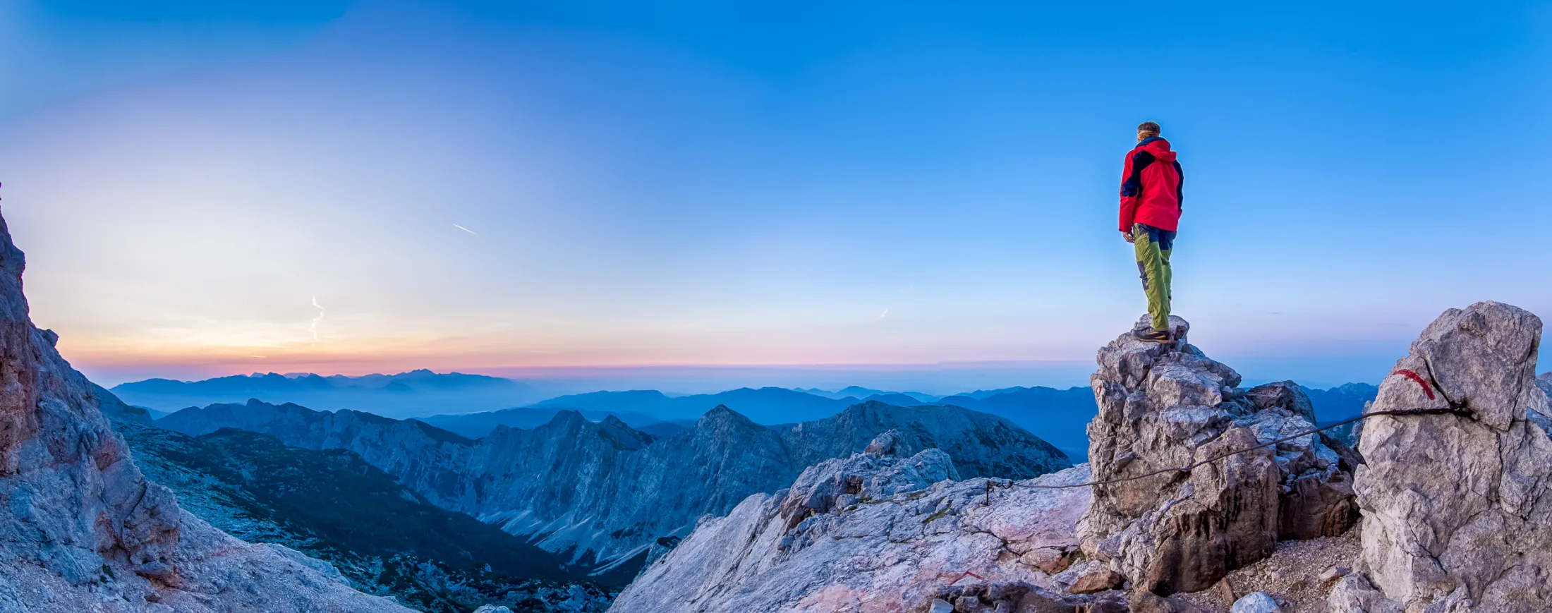 A hiker in a red jacket stands on a rocky peak, looking out over a vast mountain landscape during a clear, blue sunrise.