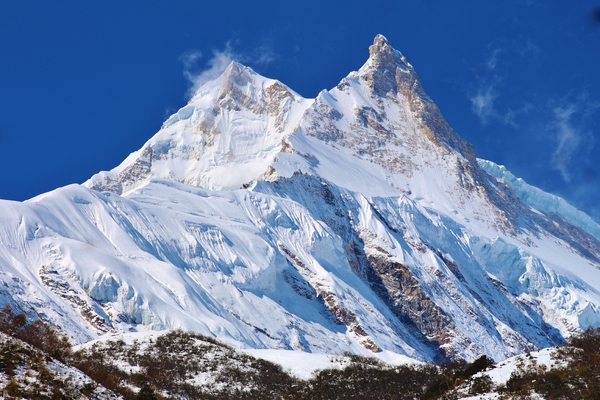 A jagged, snow-covered mountain peak with steep icy slopes rises sharply against a clear blue sky.